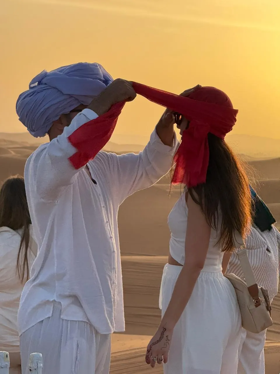 Guide helping a traveler tie a desert scarf beside the dunes