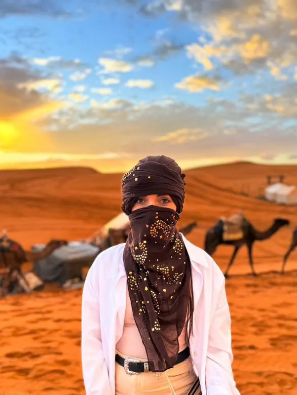 Viajero con un pañuelo oscuro del desierto, con camellos y las dunas de Merzouga detrás al atardecer