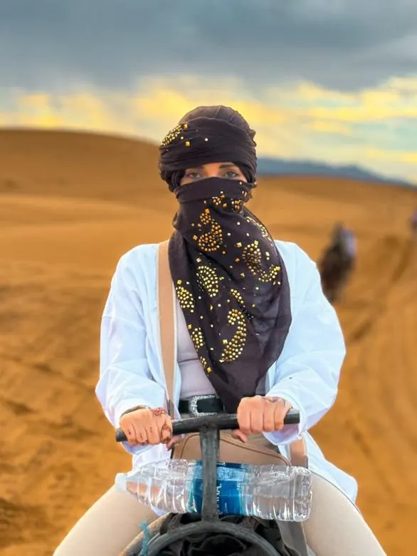 Viajero con camisa blanca y pañuelo oscuro montando en camello por las dunas de Merzouga